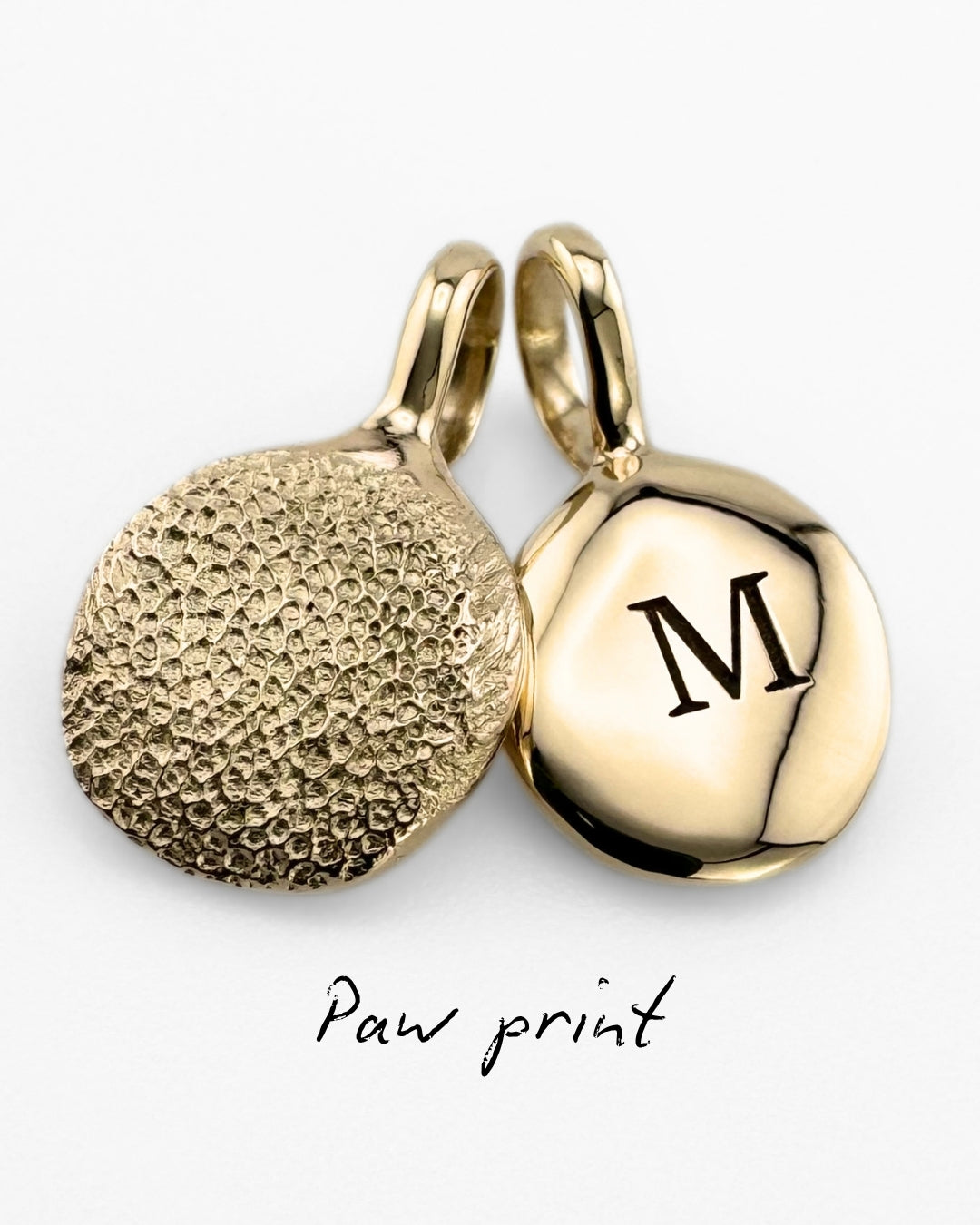 Close up of two gold pendant with dog's paw print impressions and 'M' engraving