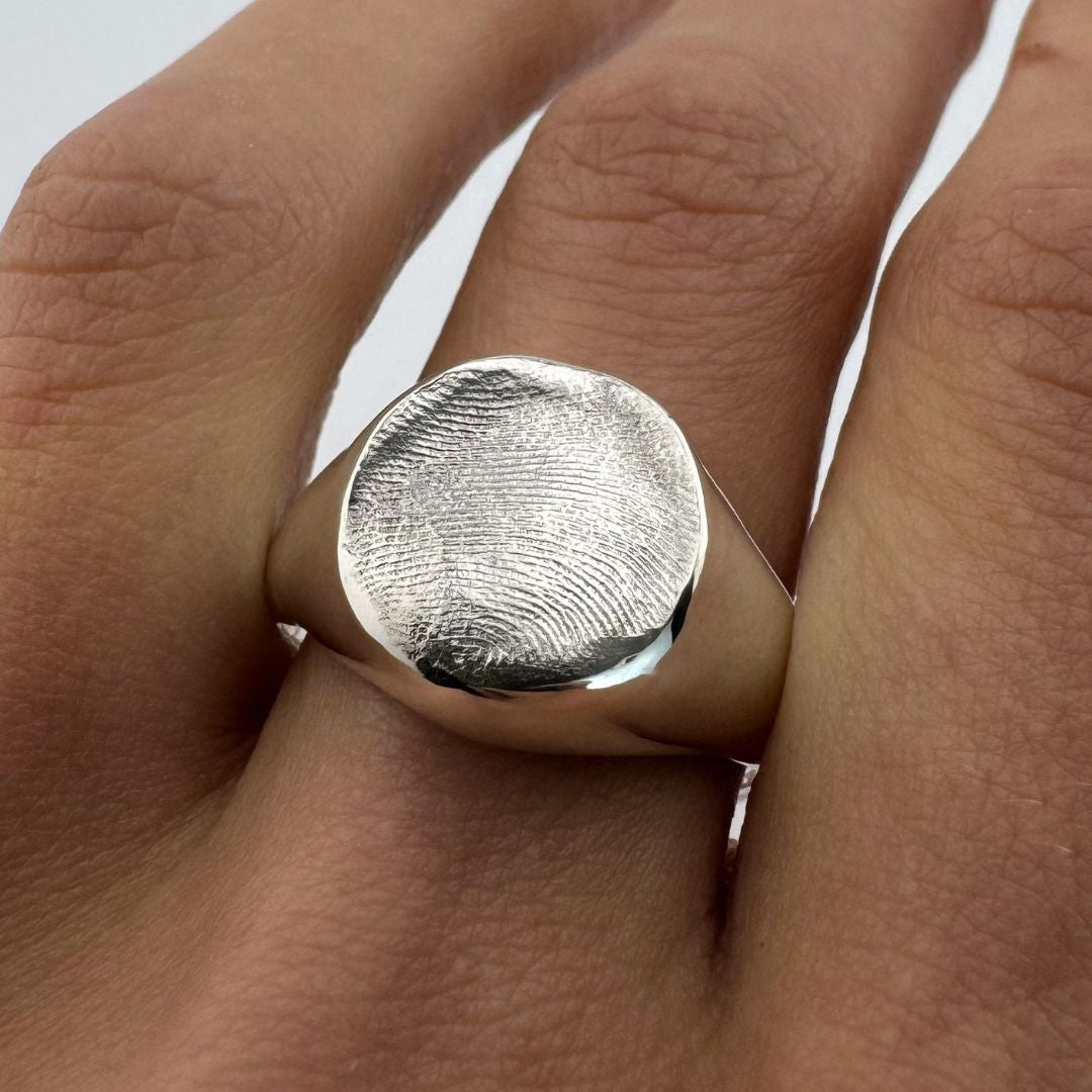 Hand wearing a large silver signet ring with fingerprint impression on a light grey background