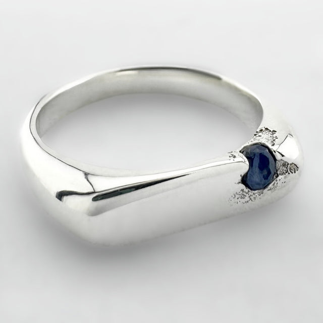 Thin silver signet ring with a blue sapphire on a light grey background